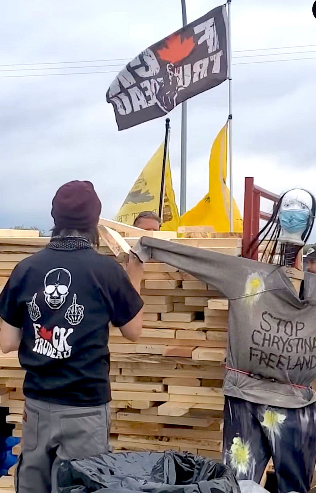 A man in a Fuck Trudeau hoodie builds a structure next to an effigy depicting Deputy Prime Minister Chrystia Freeland. A Fuck Trudeau flag flies above.