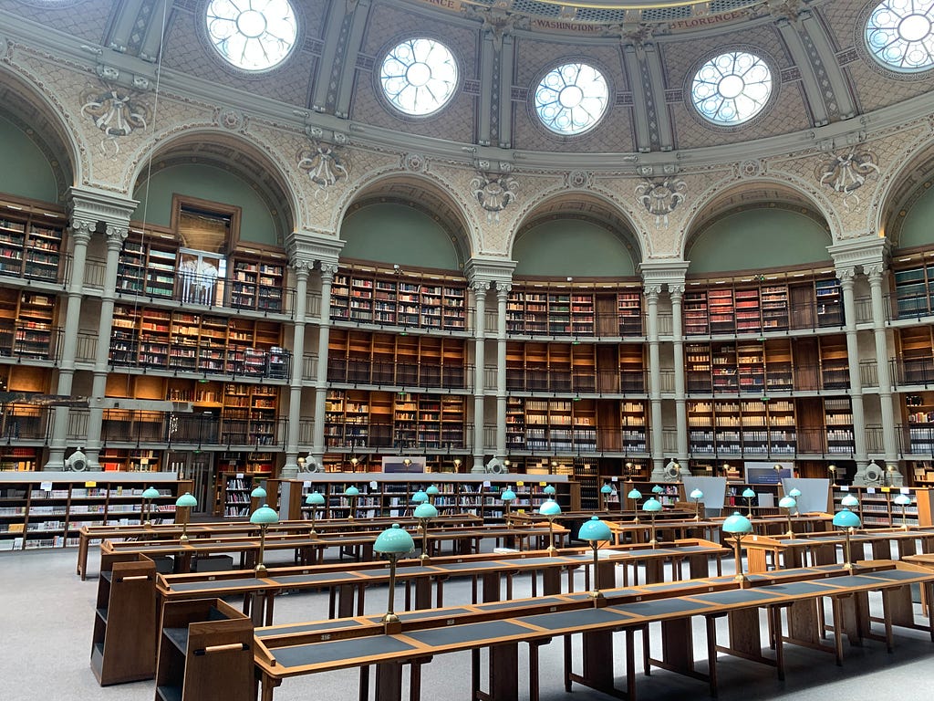 Vast BNF Richelieu library with vaulted ceiling, ornate arches and three levels of shelves overflowing with books. Reading tables with green lamps are neatly arranged, inviting study and reflection.