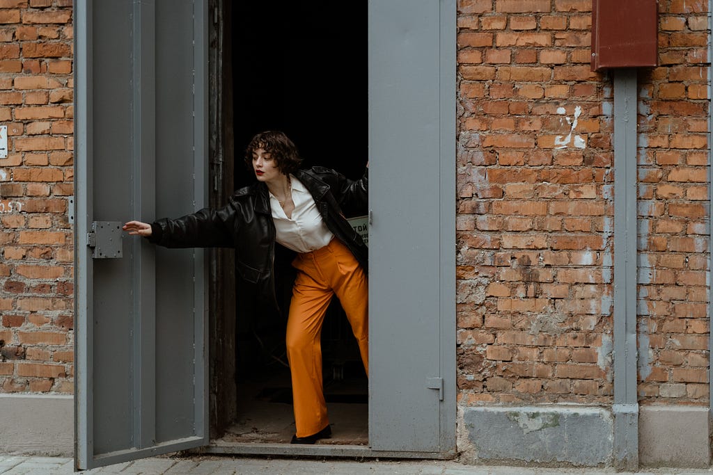 A woman in a black leather jacket, white top, and orange pants is closing a metal door on a brick building.
