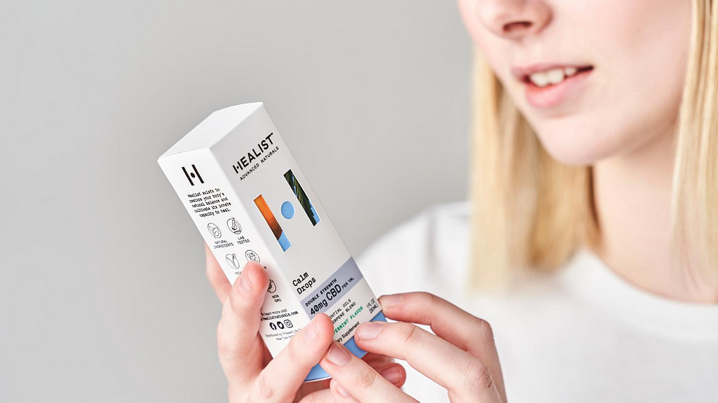 A smiling white woman with straight blonde hair examines a box of Healist Naturals Calm Drops.