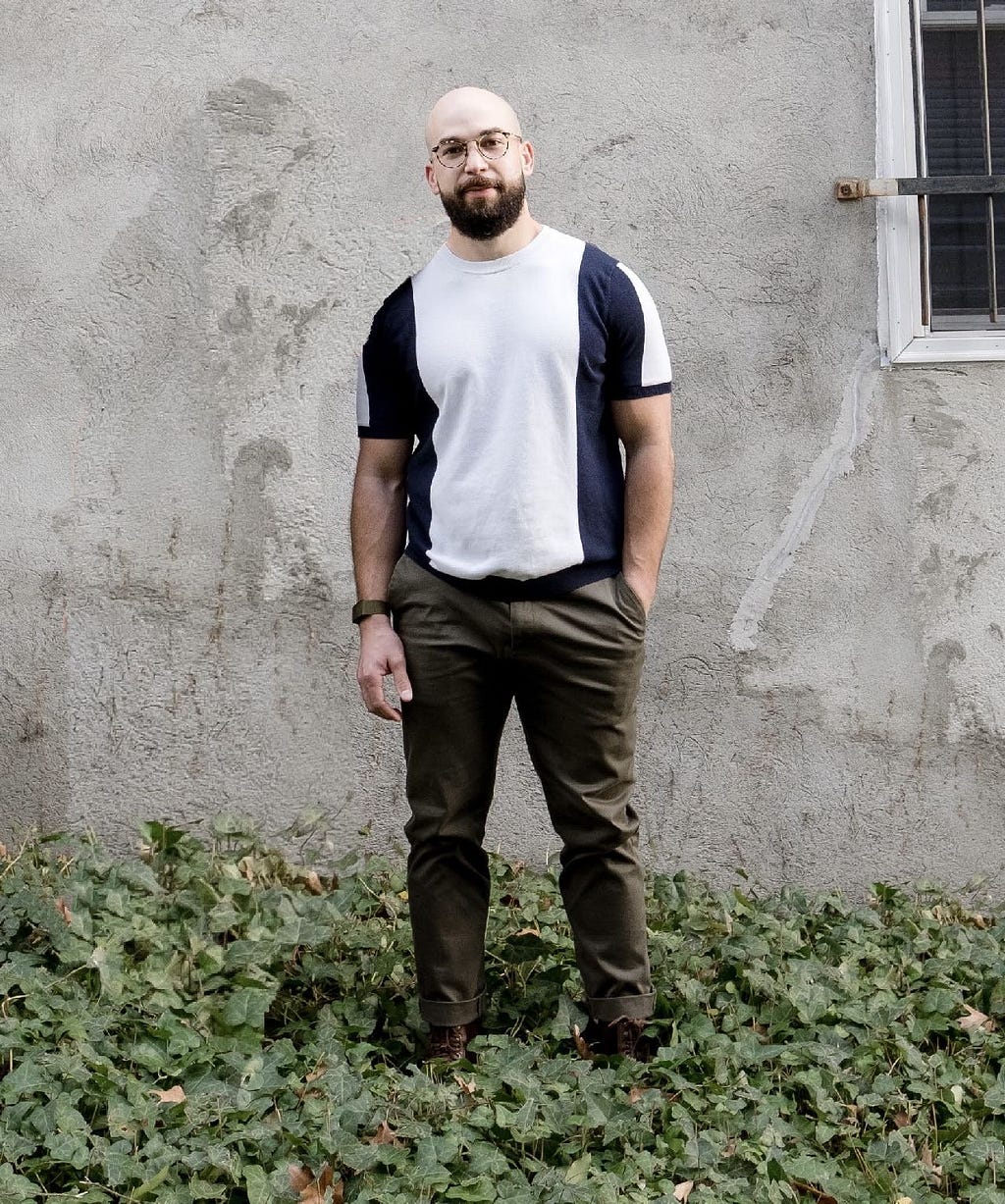 Félix stands outside on a sunny day, in ankle high greenery, in front of a gray wall. He is bald, with round glasses and a beard. He is wearing a navy and white striped shirt and army green pants, with his left hand in his pocket.