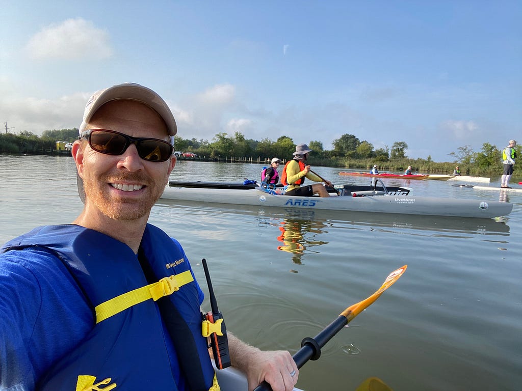 In a selfie photograph, Joel Dunn, wearing blue with sunglasses and a baseball cap, faces the camera while sitting in a kayak in the water. The kayak isn’t visible but the paddle in his left hand is. Other kayakers are visible behind him.