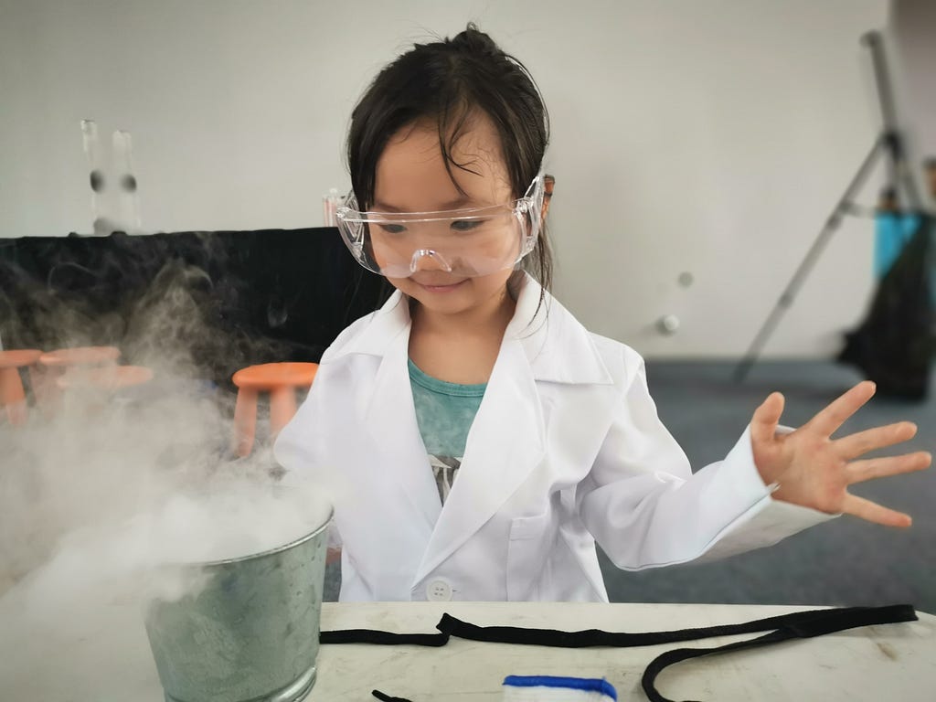 Girl of Asian descent, about 6 years old, wears a white lab coat and goggles with her arms spread and her fingers open. In front of her is a bucket with steam coming out of it. She appears to be doing a science experiment.