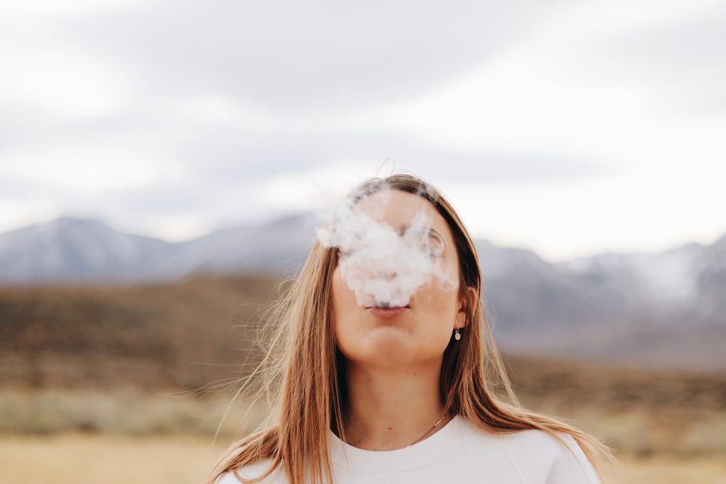 Woman blows a puff of smoke into the air while standing in front of a mountain landscape.
