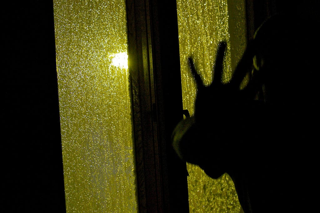 A shadowy hand is held in front of the camera with a backdrop of a window with a streetlight filtering through.