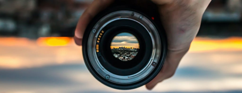 Close up of hand holding a camera lens, with view of distant city in focus.