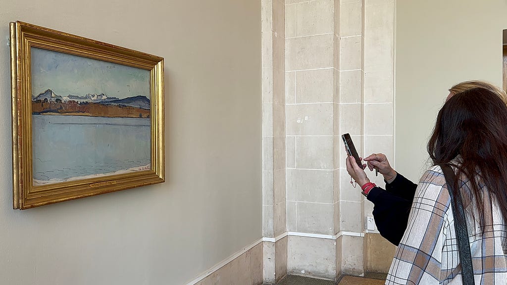 Two persons photographing a framed landscape painting in an art gallery. The artwork depicts a serene lake with mountains in the background, and is hung on a pale wall next to a tall window.