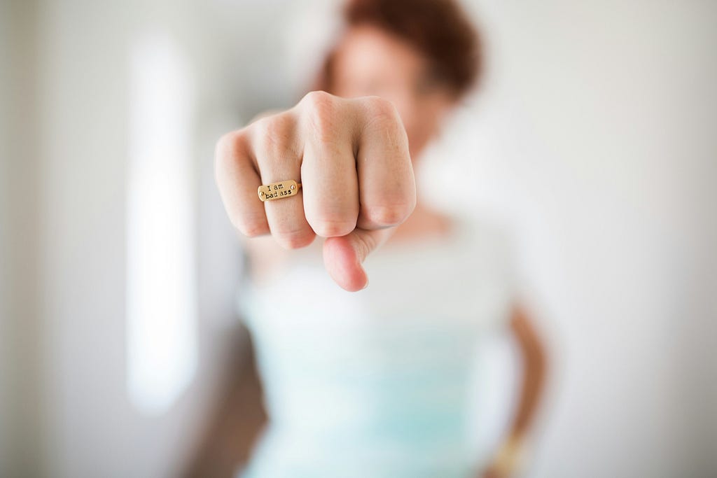 A woman extending her fist toward the camera wearing a gold ring that says I am bad ass.