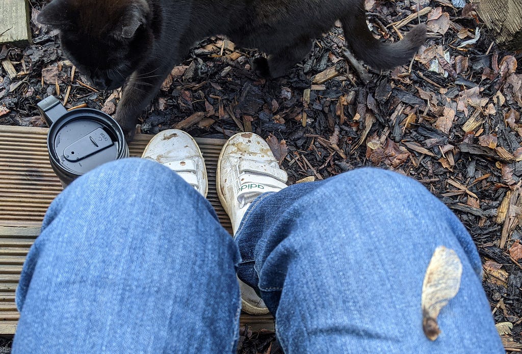 A photo of me — looking down at my legs, with a hot drink and Pepper black cat close by