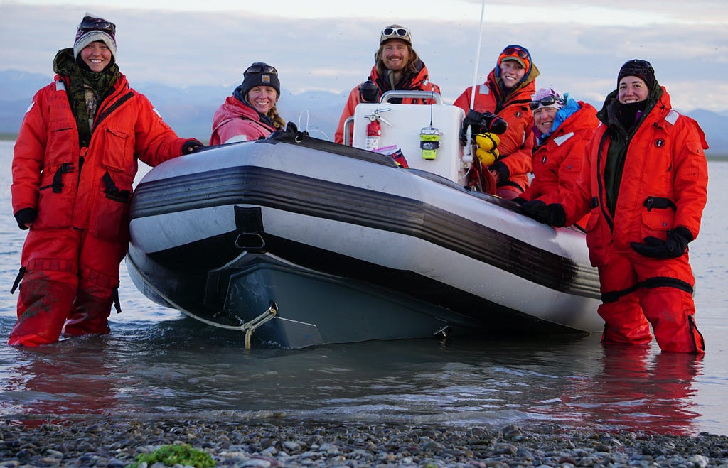 A group of people in orange mustang survival suits around an ocean inflatable skiff.