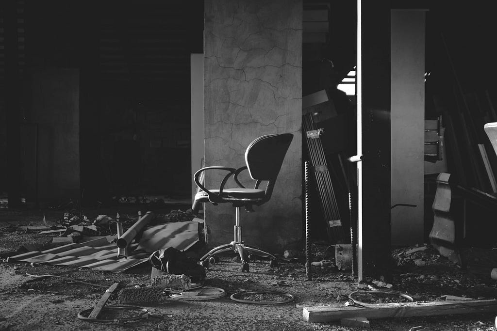 An office chair is in the middle of an empty room, with debris and broken materials surrounding it. All is in black and white, with dark shadows in the background.