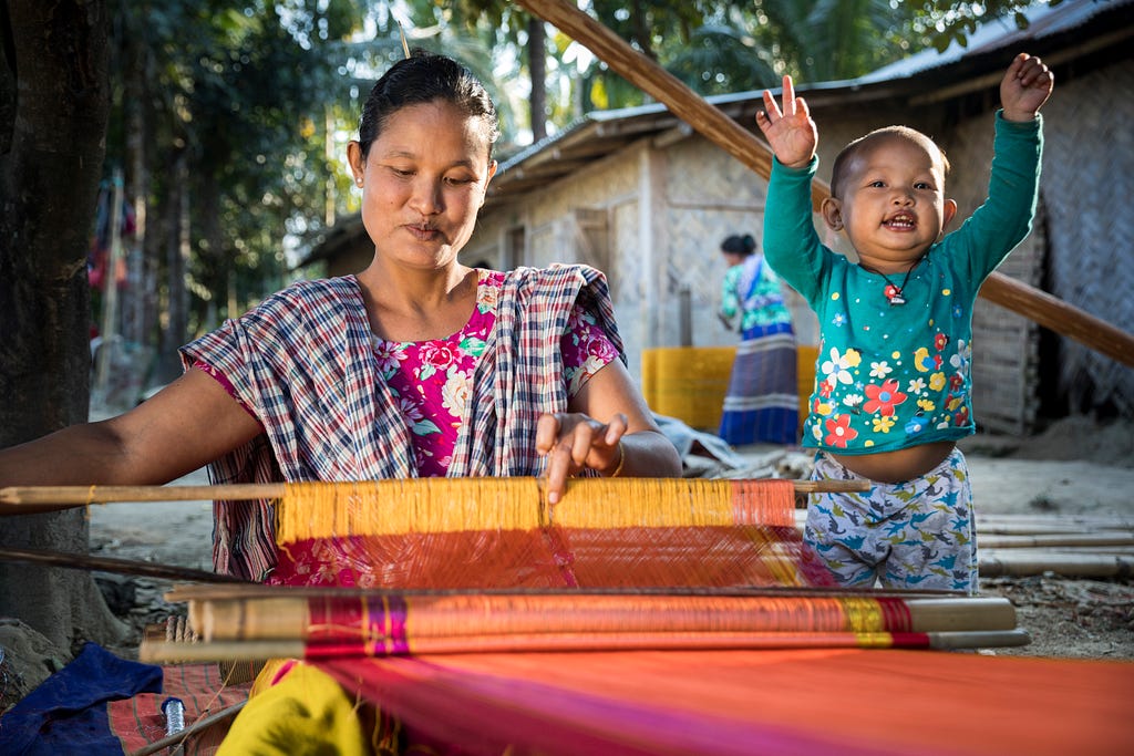 A woman works on a loom with vibrant orange and pink fabric while a small child beside her raises both arms in the air.