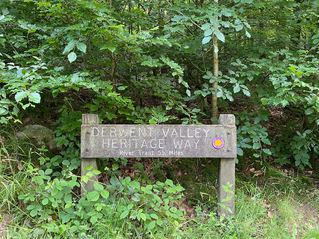 A wooden sign reads ‘Derwent Valley Heritage Way River Trent 55 miles’. Behind are green foilage and trees.