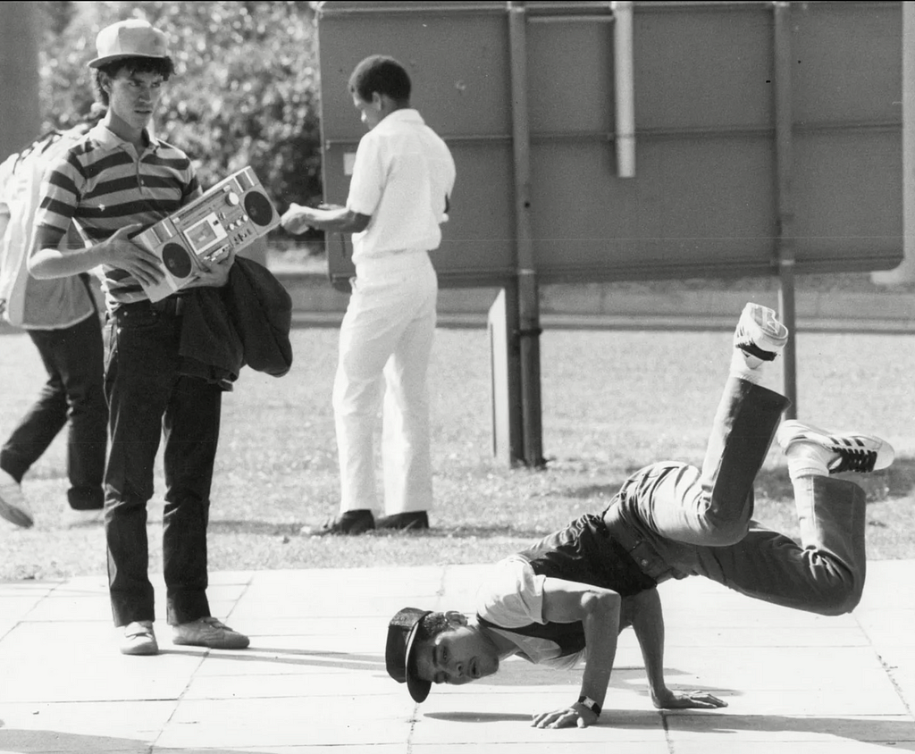 A bboy is practicing his moves alone on pavement, as a man standing on the side holding a boombox fixates him.