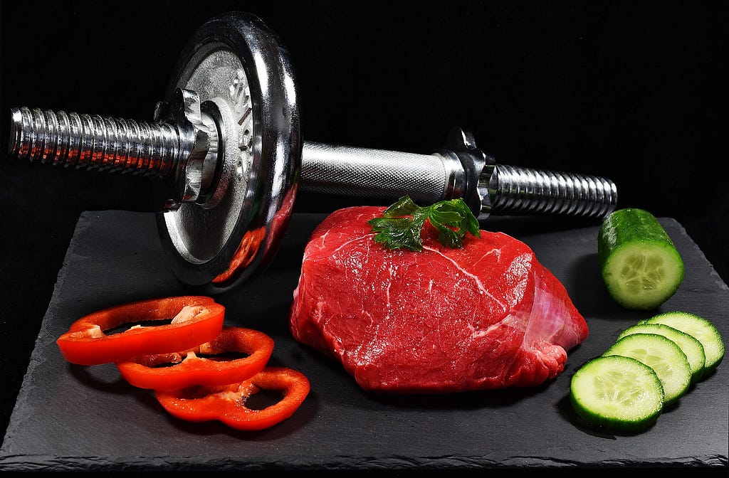 A dumbbell with one plate missing sits diagonally behind a slab of raw beef, sliced red bell pepper, and cucumber.