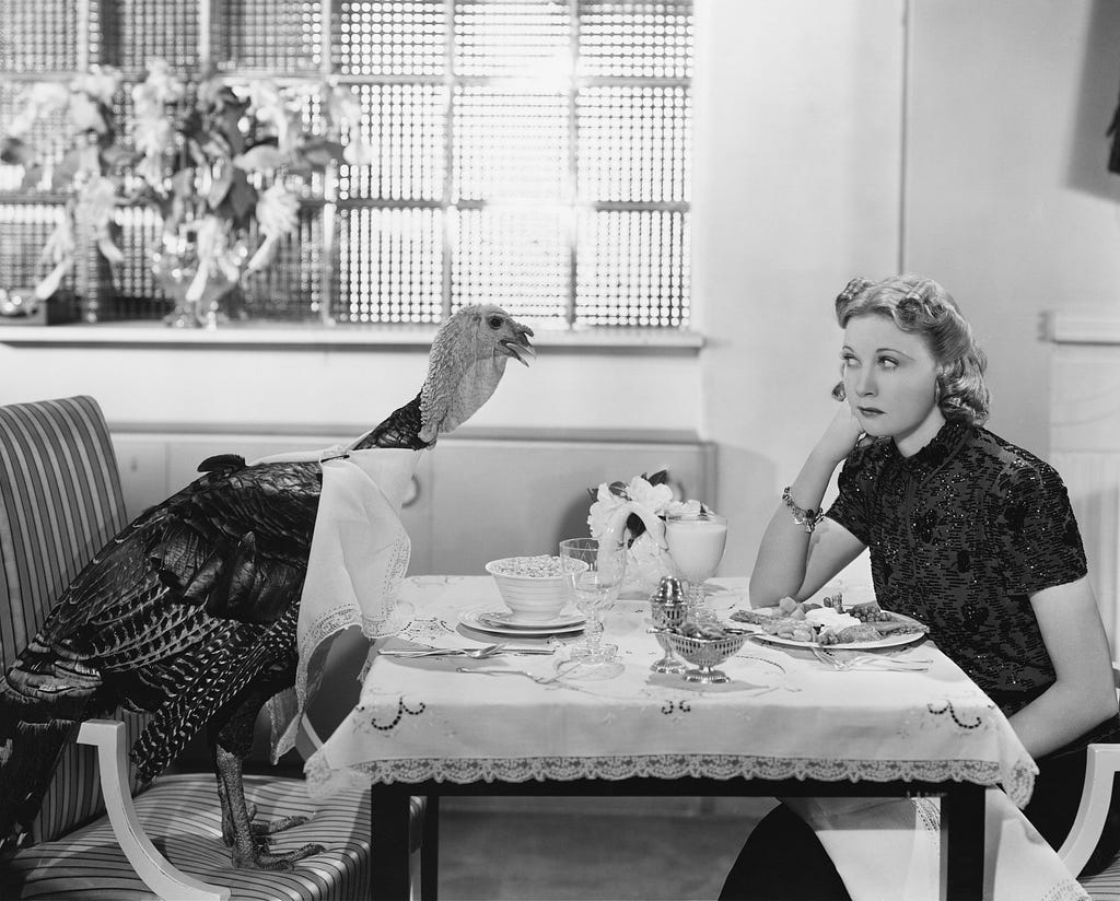 A woman sitting at a table with a turkey sitting across from her on Thanksgiving