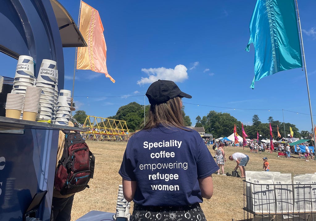 A woman with long brown hair and a black cap at a festival, wearing a tshirt that reads ‘Speciality coffee empowering refugee women’