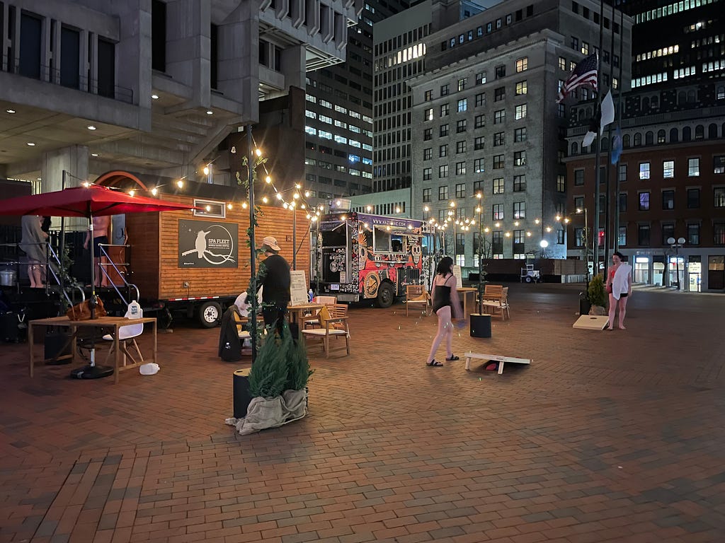 The mobile pop-up sauna at night. String lights surround the sauna and adjacent seating area with several people seated, standing, and playing a game of corn hole. A food truck offers Mexican food for sale.