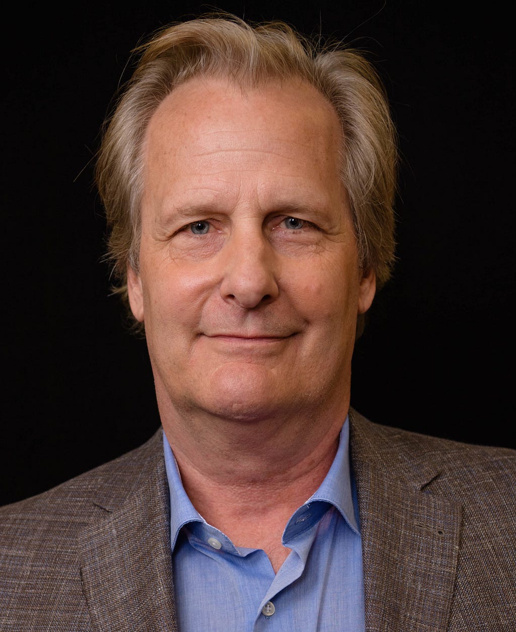 Picture from the shoulders on up of the actor Jeff Daniels, a white male in his fifties with blonde-gray hair and blue eyes. He’s sporting a closed-lipped smile and a blue shirt and brown sports jacket.