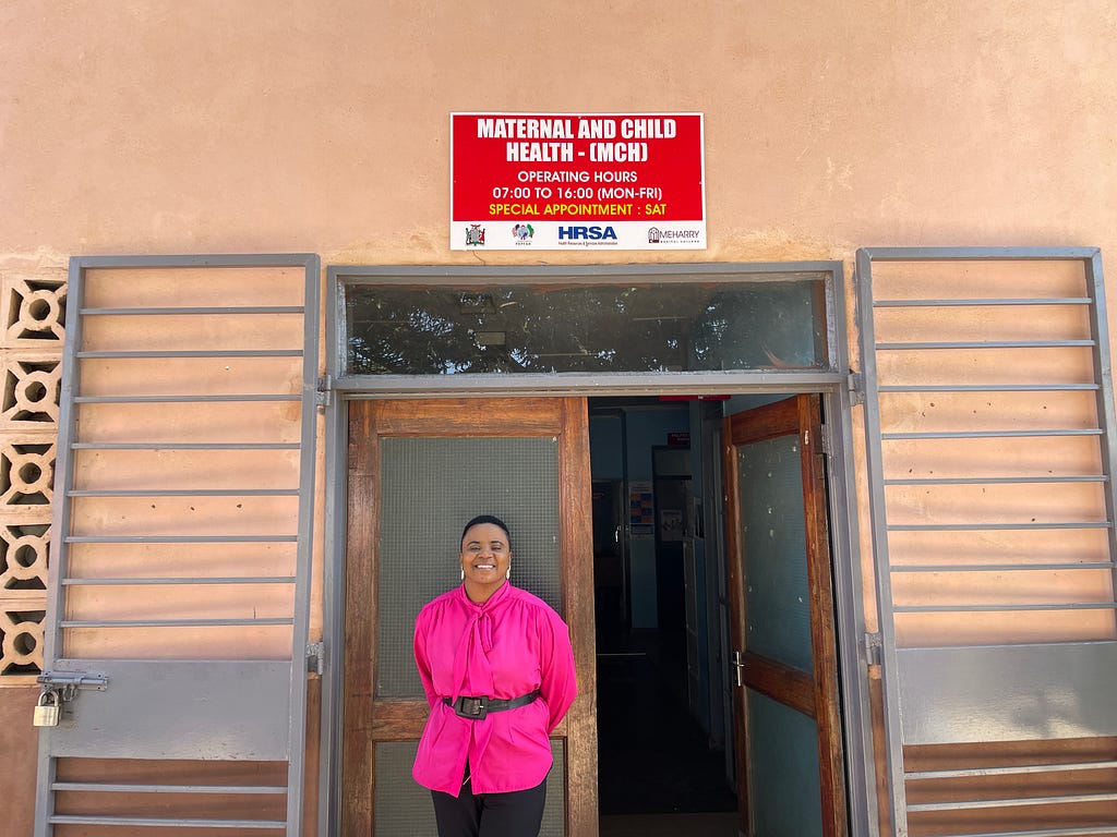 Esnart is wearing a bright pink blouse, and is standing outside of the maternal and child health ward, which is a beige pink building with a bright red sign.