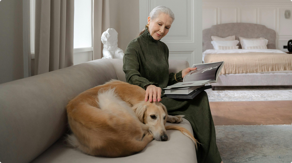 An older woman sitting on a sofa reading a book, with a large dog resting beside her.