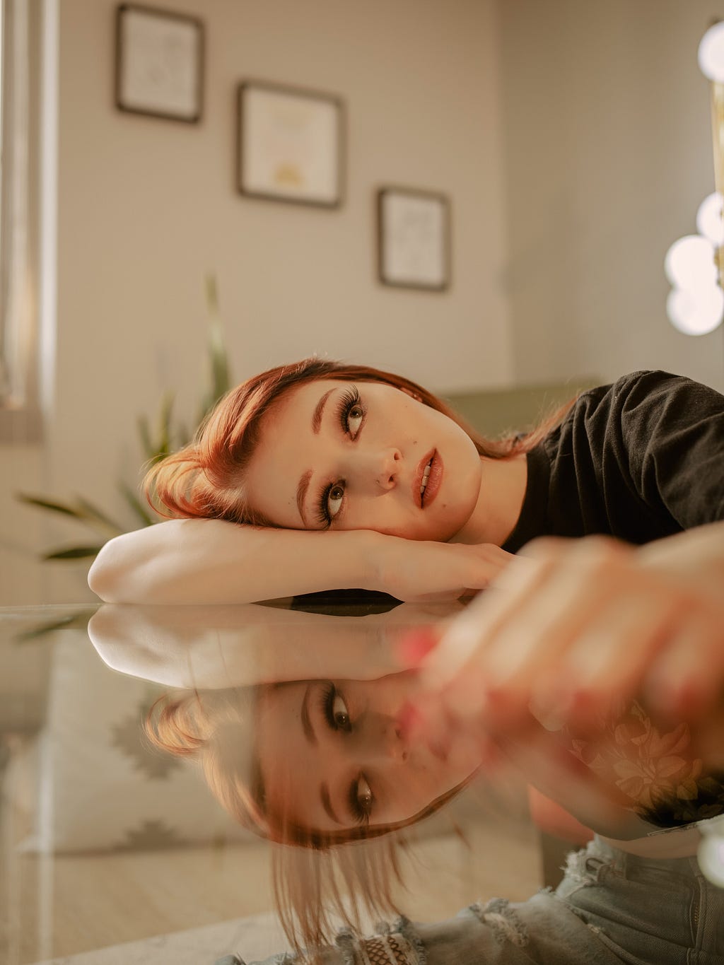 A red headed woman laying her head down on her arm on a glass table top reflecting upon something, as her image reflects back on herself.
