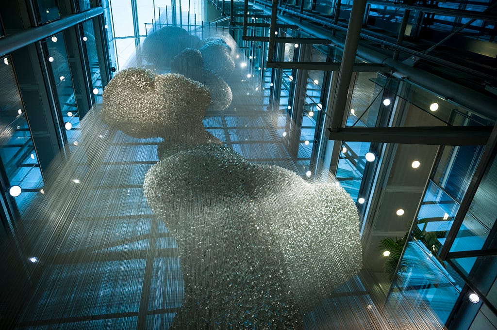 A photograph looking up from the ground floor of a large (multi-story) sculpture. The sculpture is a sinuous structure constructed of thousands of small glass balls strung along hundreds of long stainless steel wires that run from floor to ceiling. The effect is a beautiful 3D curving shape inspired by the shape of molten lead dropped into water forms.