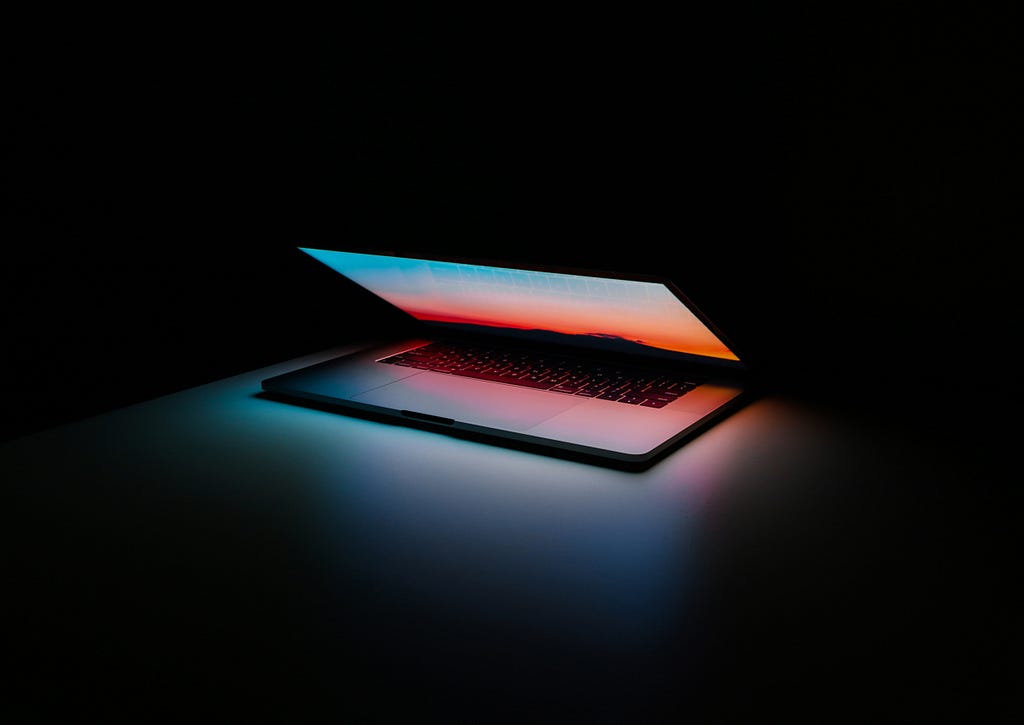 A laptop sitting on a desk in a completely darkened room, the lid of the device is just ajar to let the light from the screen shine out in colours of blue and orange.