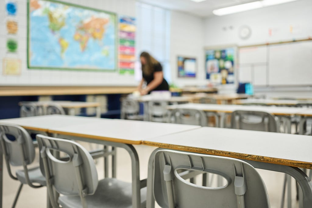 Photo of a classroom with desks and chairs in the foreground and a teacher in the blurred background. Photo by AJ_Watt/Getty Images