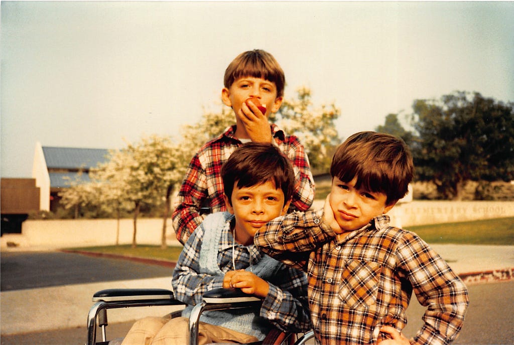 Description: This image shows three brothers wearing plaid shirts. The boy in the middle sits in a wheelchair in front of Roy