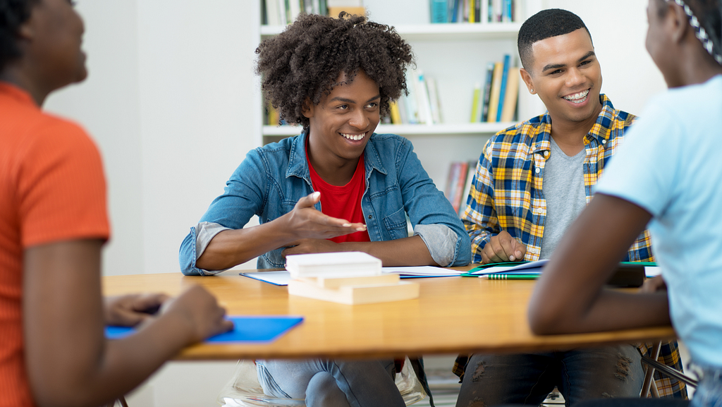 Group of four Black students sit around a table in front of a bookshelf. Two students face the camera in discussion with another student, their faces look happy and engaged.