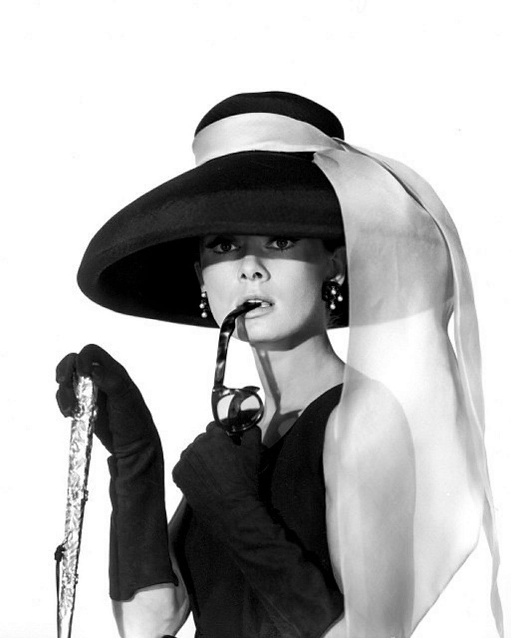 Headshot of Audrey Hepburn. She is wearing long black gloves, a black dress, and a black hat with a light-colored scarf.