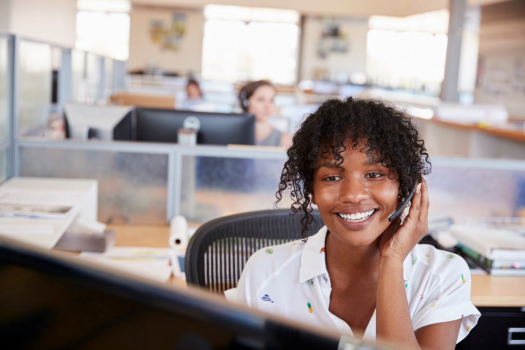 A woman sits at a desk, wearing a headset and smiling at her computer.