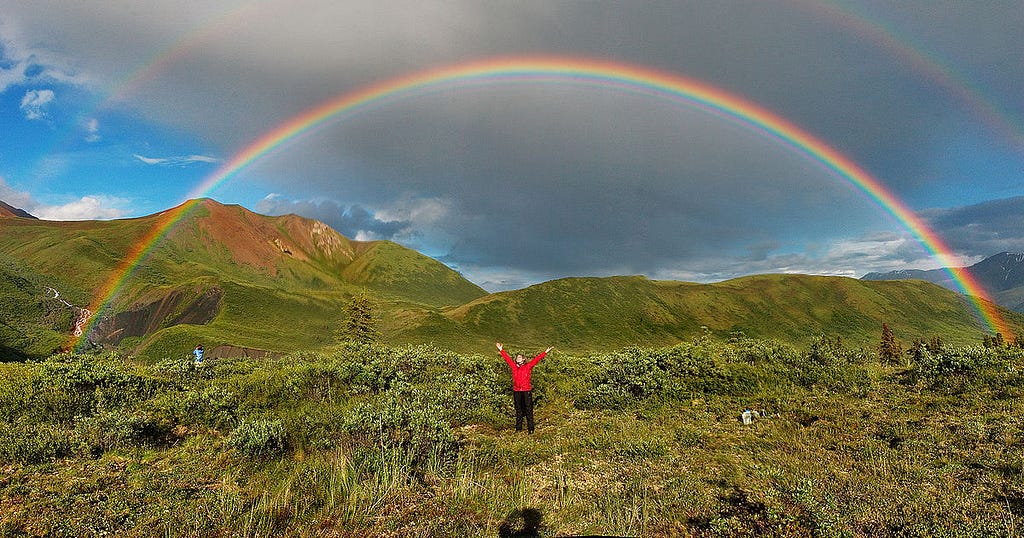 A photograph of a double rainbow over a green pasture. A figure is stood underneath.