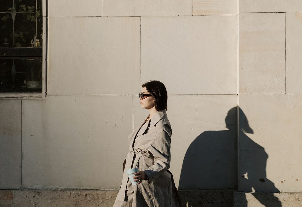 Photo of a woman walking in the sidewalk wearing a trench coat, sunglasses, coffee in hand, and looking cool!