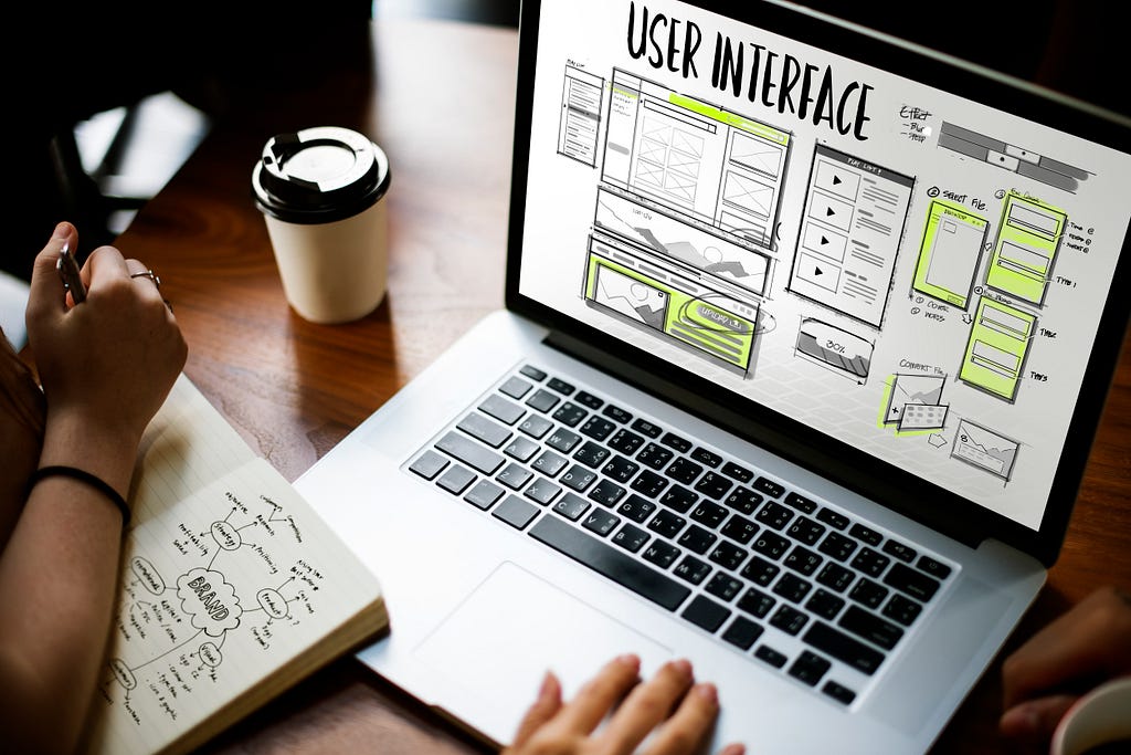 Fotografia de uma mesa de escritório com um notebook caderno e copo de café. Na tela do notebook podemos ver wireframes em preto e amarelo com o título “user interface” em destaque.