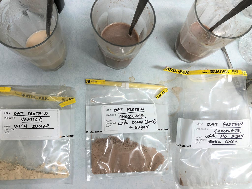 Three cups of water mixed with Upright Instant Oatmilk next to packaging with labels for taste testing. From left to right: vanilla with sugar, chocolate with sugar, and chocolate with no sugar.