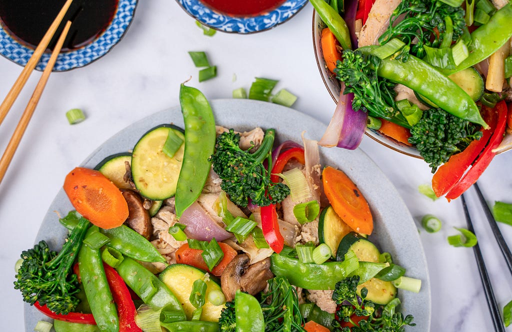 A top-down view of a plate and a bowl of stir fry with chicken, broccolini, carrots, zucchini, red peppers, mushrooms, and green onions.