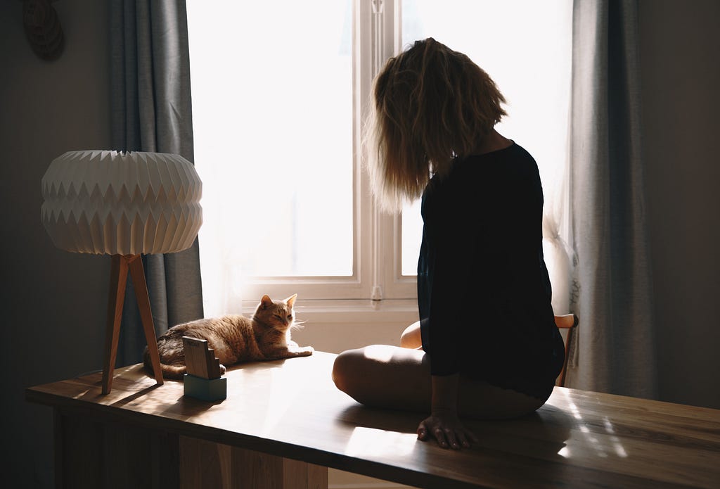 A girl sits on a table looking out the window with her orange cat beside her looking at her. Her back is to the viewer.