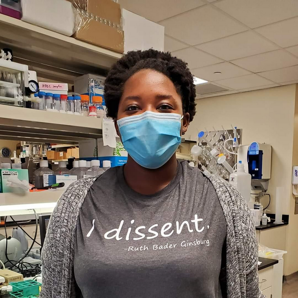 Ariele, a black woman wearing a grey shirt with the text “I dissent- Ruth Bader Ginsburg”. She is wearing a mask, standing in a laboratory