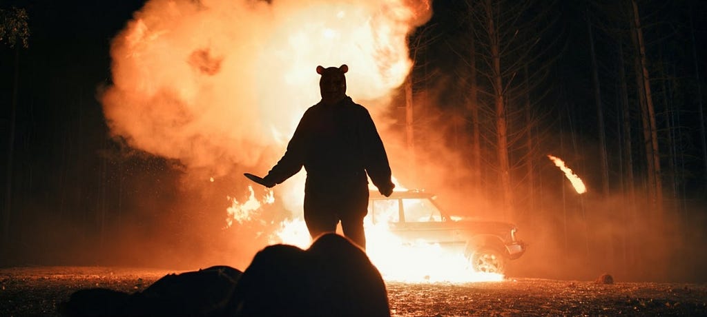 A film still of a monstrous Winnie the Pooh in silhouette, holding a knife, stood in front of an exploding car