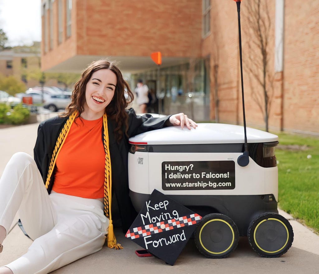 Taylor, in a graduation gown and orange shirt, sitting behind a Starship Delivery Robot posing for a photo with her grad cap decorated to say, “Keep Moving Forward”