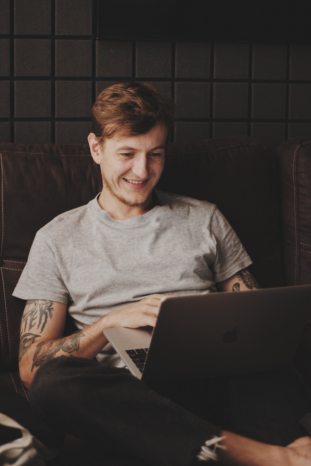 A young man sits and smiles while using his laptop.