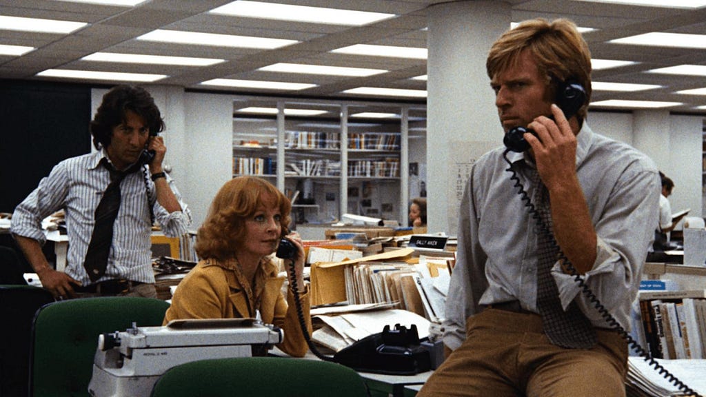 Still from the movie All the President’s Men. Two men and one woman in a newsroom, all on the telephone.