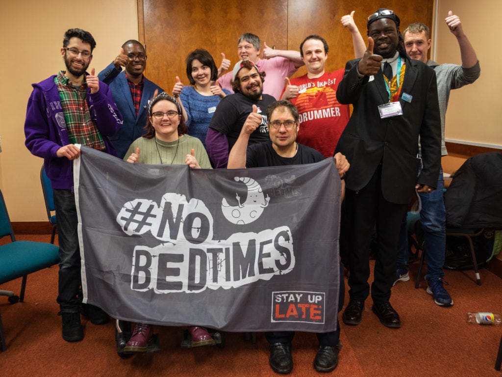 Group of campaigners with learning disabilities holding a flag that says ‘No Bedtimes’. They are all smiling and holding their thumbs up.