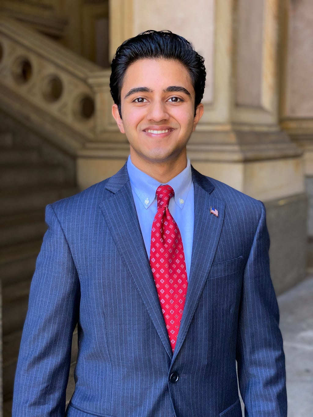 A young man wearing a pinstriped blue suit with a red tie and American flag pin poses with a smile for a picture in City Hall.