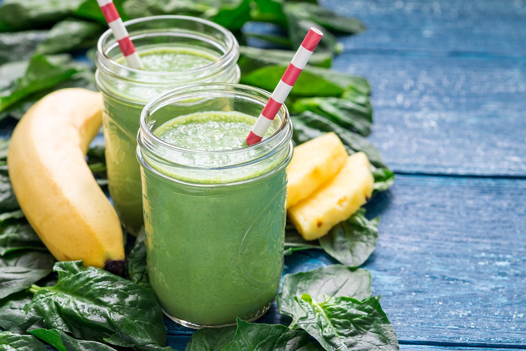 2 jars filled with a green smoothie, surrounded by spinach leaves, pineapple chunks and a banana.