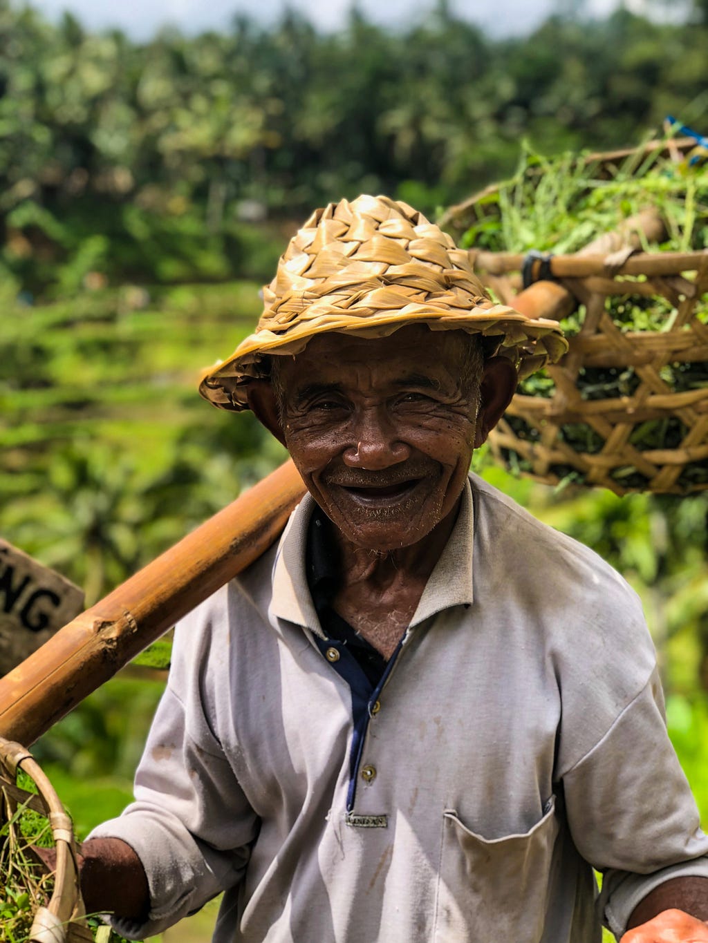 Older man smiles at the camera with a basket of vegetables on his shoulder