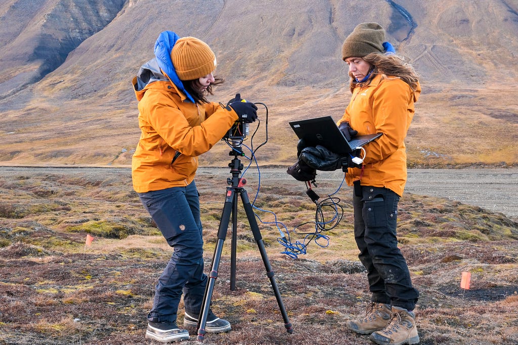 Jess Todd and Cody Paige wearing orange winter jackets and setting up a LiDAR camera on a tripod at the base of a brown and green mountain.