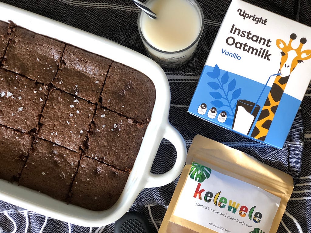 Vegan Kelewele plantain brownies in a white glass brownie dish with Upright Instant Oatmilk on the side in a clear glass with a spoon. Packaging for Kelewele brownies and Upright Instant Oatmilk placed alongside dishes on table.